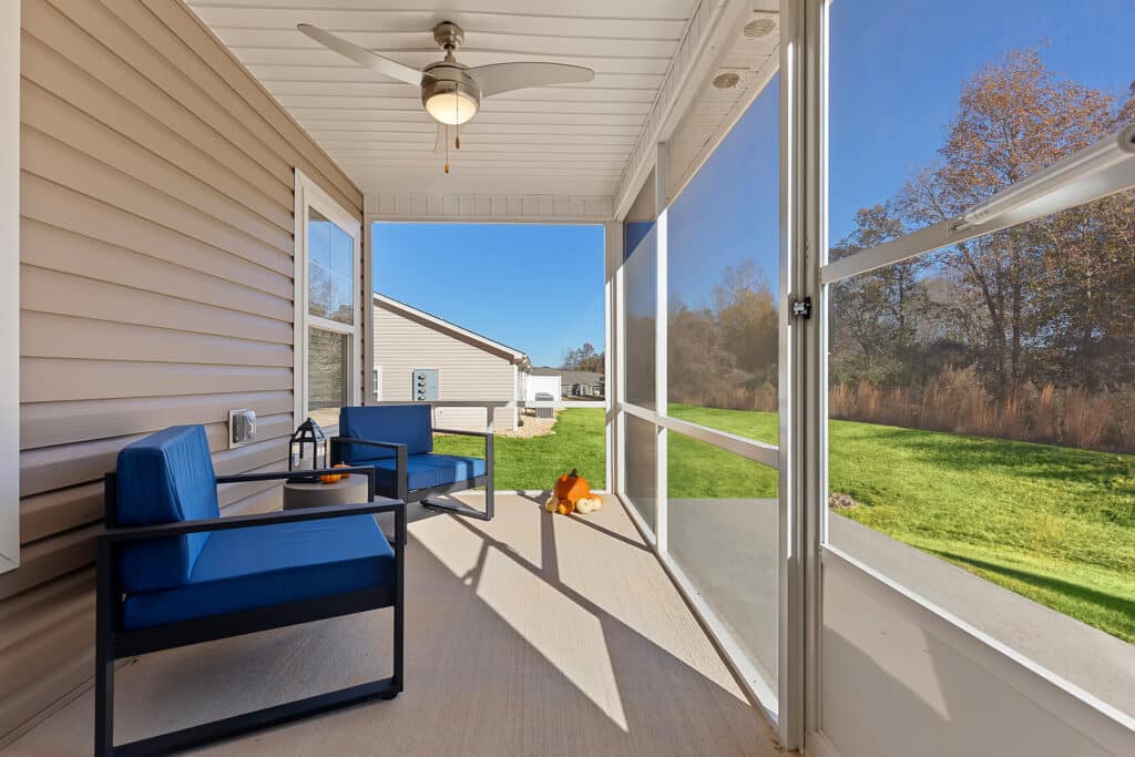 Screened-in porch with two blue cushioned chairs, a ceiling fan, and a view of a grassy yard with trees under a clear blue sky.