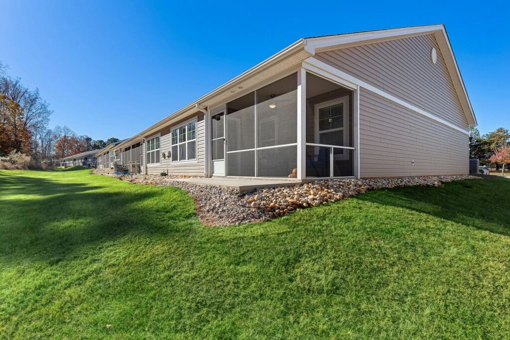 Single-story building with beige siding, screened porch, and stone landscaping set on a sloped grassy yard under a clear blue sky.