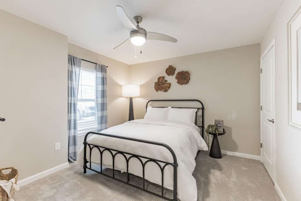A small bedroom with a metal bed frame, white bedding, a ceiling fan, a floor lamp, two doors, and a window with plaid curtains. Decorative wall hangings are above the bed.