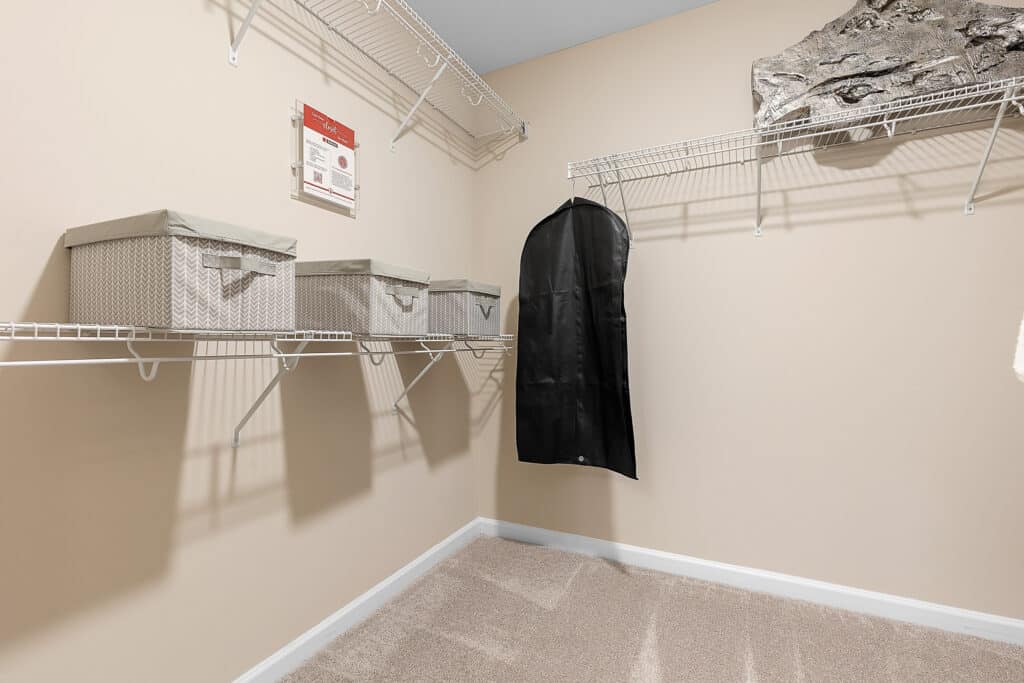 Empty walk-in closet with beige walls, wire shelves holding three fabric storage boxes, a black garment bag, and a duct covered by insulation in the corner.