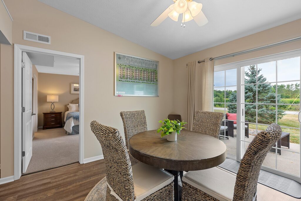A dining area with a round table and four woven chairs, a potted plant centerpiece, sliding glass doors, and a view into a bedroom.