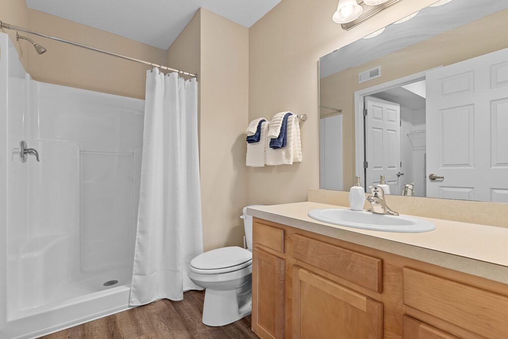 A bathroom with a shower and white curtain, toilet, wooden vanity with sink, large mirror, and folded towels on the wall.