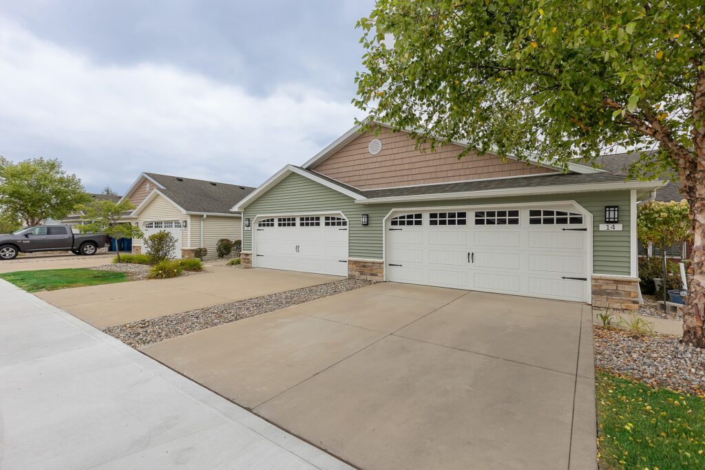 Duplex house with two white double garages, beige siding, concrete driveways, and a small tree on the right side.