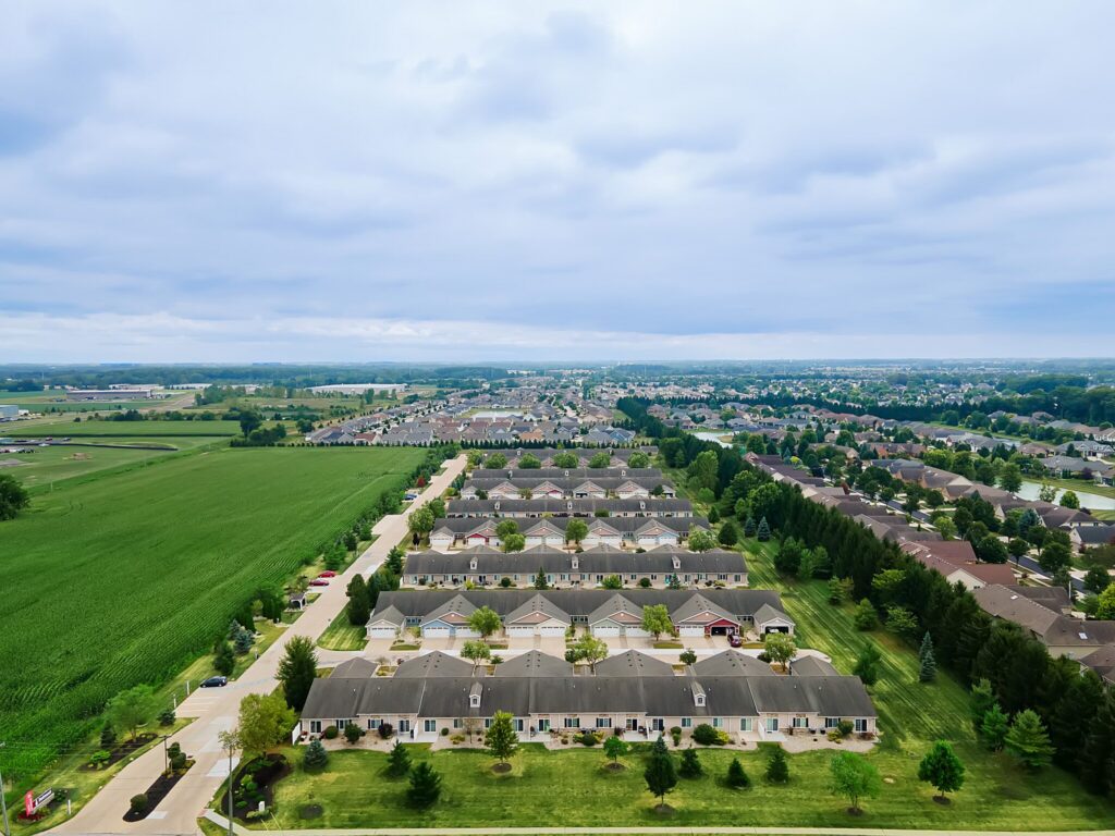 Aerial view of a suburban neighborhood with rows of houses, green lawns, surrounding trees, and adjacent farmland under a cloudy sky.
