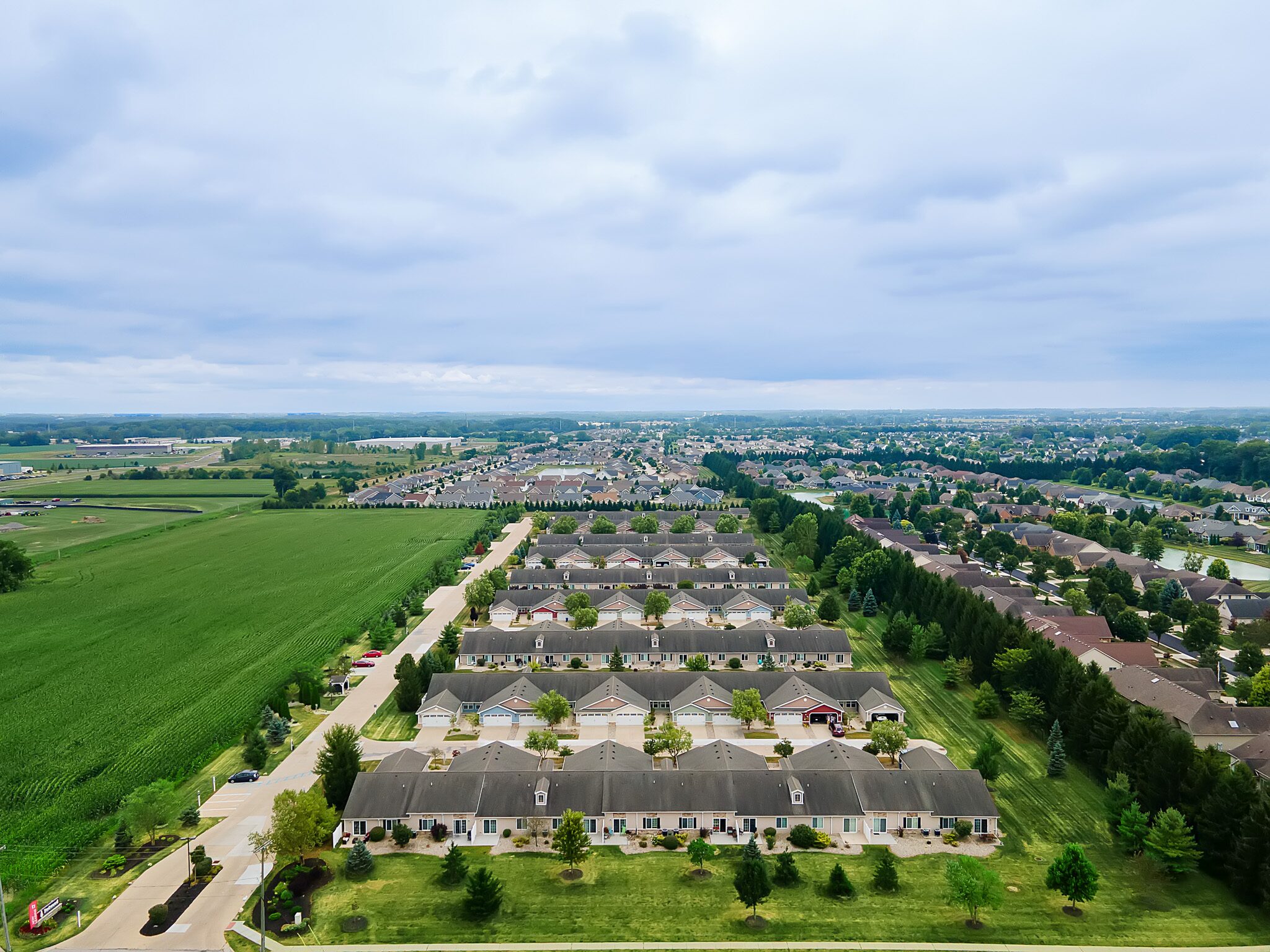 Aerial view of a suburban neighborhood with rows of houses, green lawns, surrounding trees, and adjacent farmland under a cloudy sky.