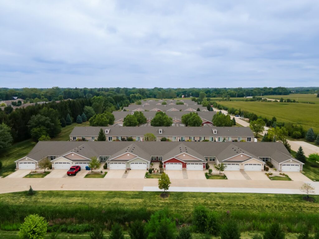Aerial view of a suburban neighborhood with rows of townhouses, garages, driveways, and green lawns surrounded by trees and fields.