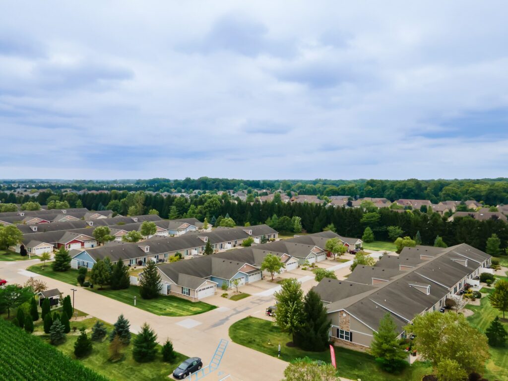 Aerial view of a suburban neighborhood with rows of single-story houses, landscaped yards, trees, and a forested area in the background under a cloudy sky.
