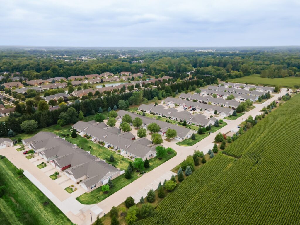 Aerial view of a suburban housing development bordered by fields and dense trees under a cloudy sky.