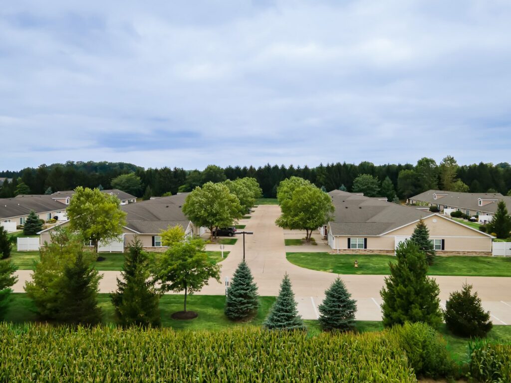 Aerial view of a suburban neighborhood with single-story houses, green lawns, trees, a central road, and a cornfield in the foreground under a cloudy sky.