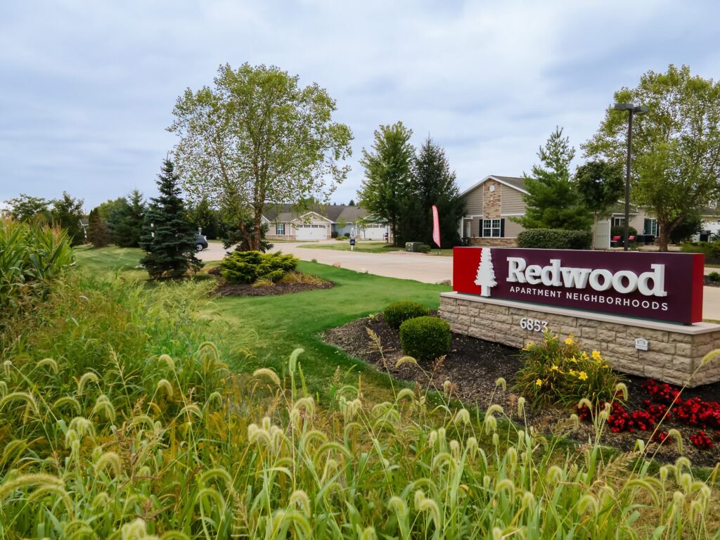 Entrance to Redwood Apartment Neighborhoods with a red and white sign, landscaped greenery, and residential buildings in the background.
