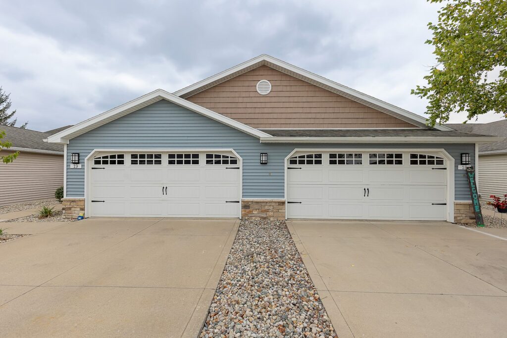 Duplex house exterior with two adjacent white garage doors, blue and beige siding, stone accents, and a rock-lined driveway divider under a cloudy sky.