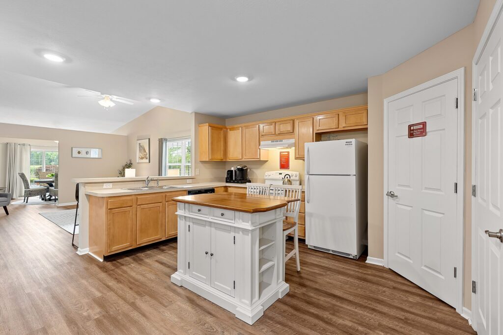 Bright kitchen with light wood cabinets, white appliances, a small island with two chairs, and wood flooring. Living area with large window visible in background.