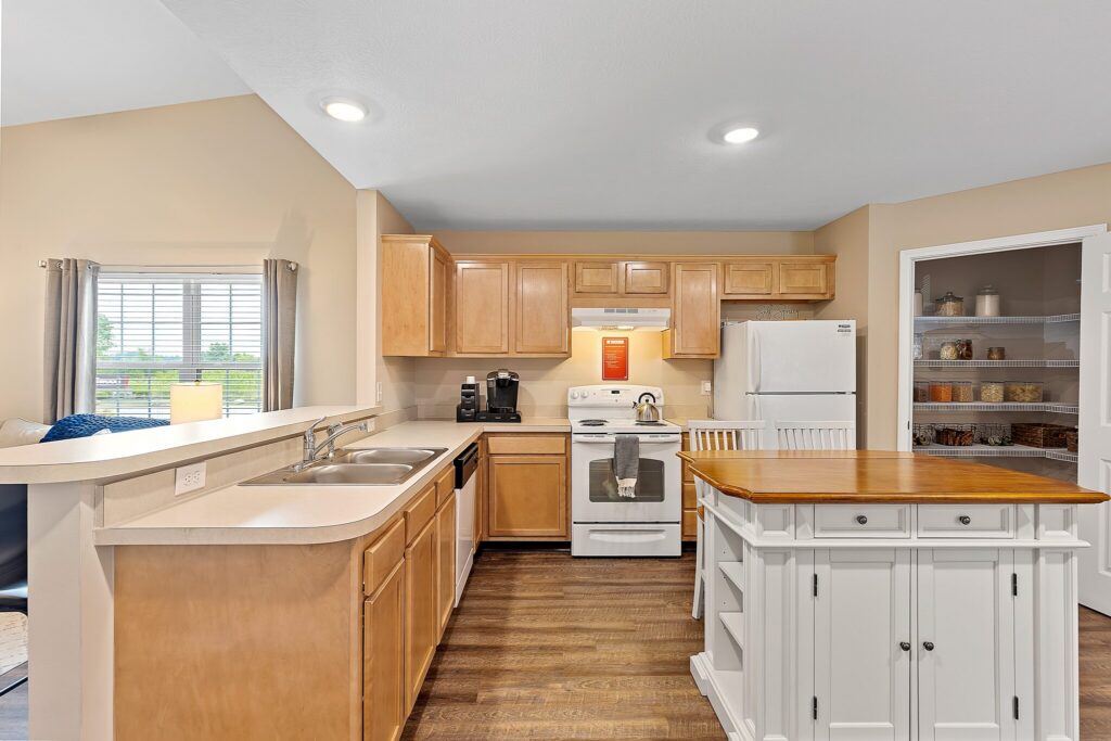 Kitchen with light wood cabinets, white appliances, a white island with wooden top, double sink, and a walk-in pantry with shelves containing jars and baskets.