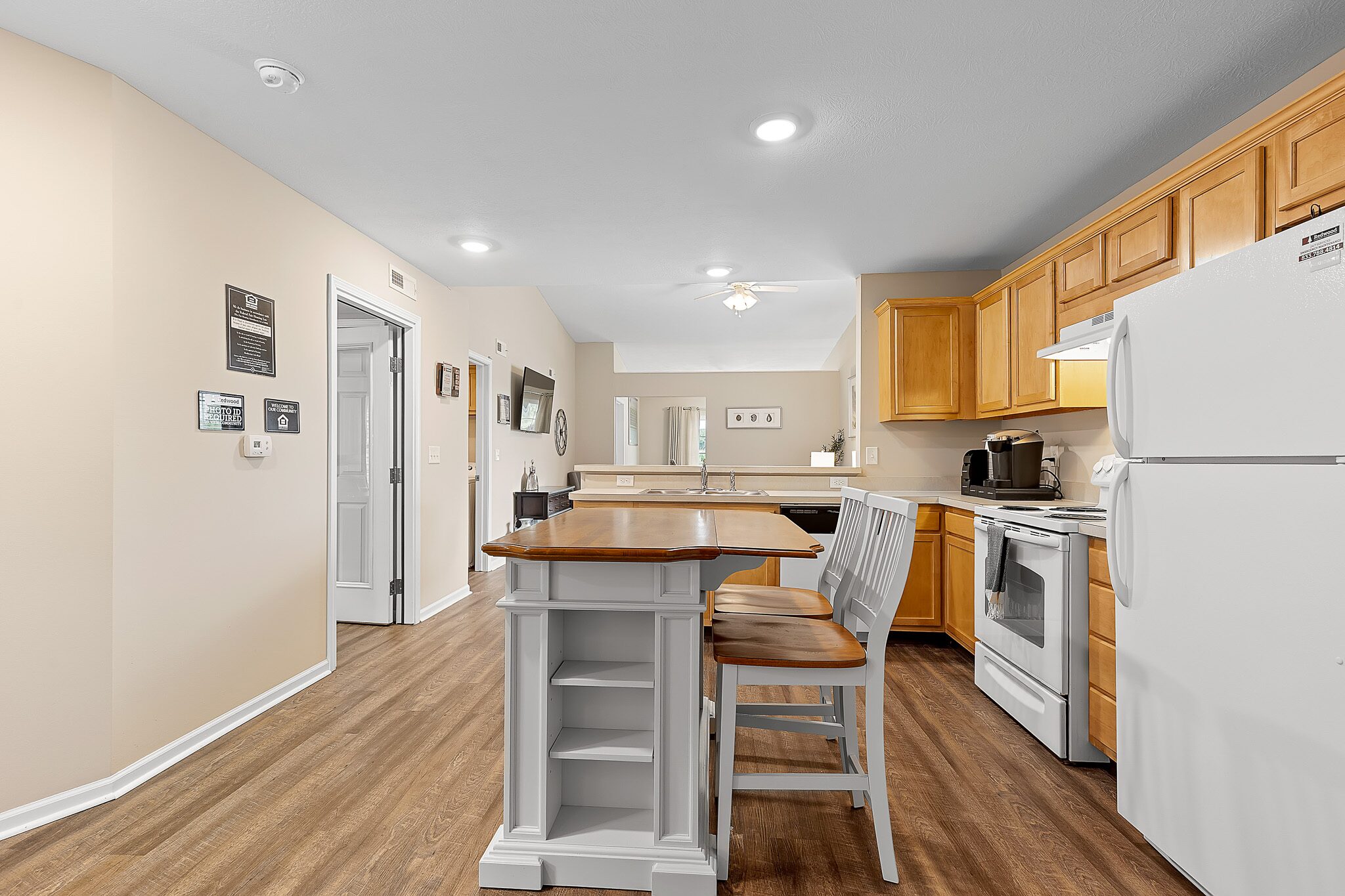 A kitchen with light wood cabinets, white appliances, an island with shelves, two chairs, and wood flooring under bright ceiling lights.