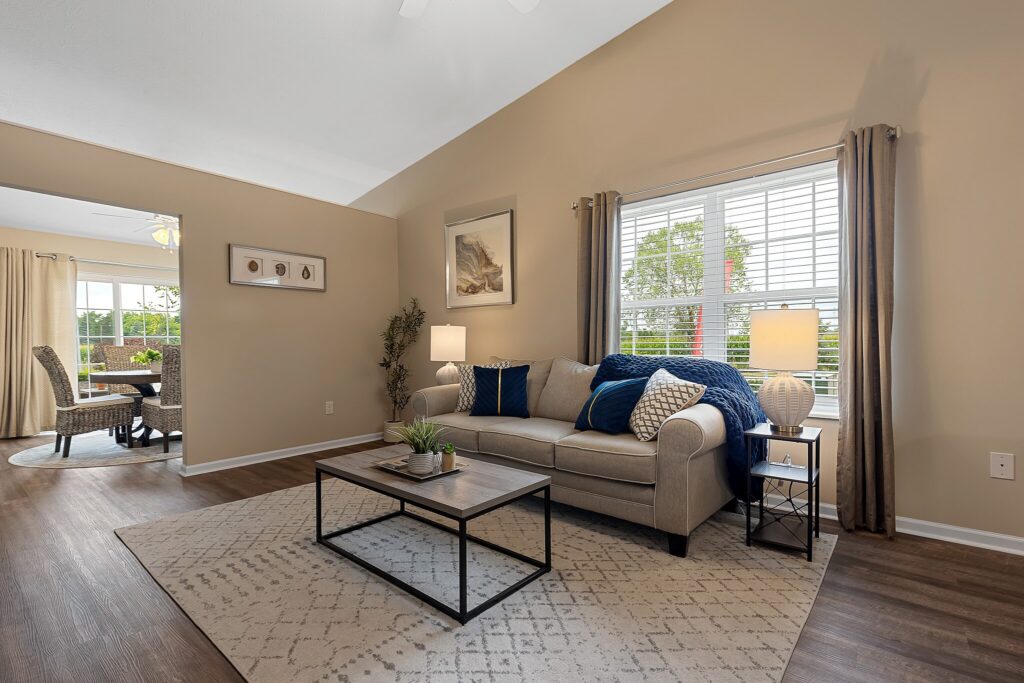 A modern living room with a beige sofa, blue pillows and throw, a wooden coffee table, lamps, and large windows with beige curtains. A dining area is visible through an open doorway.