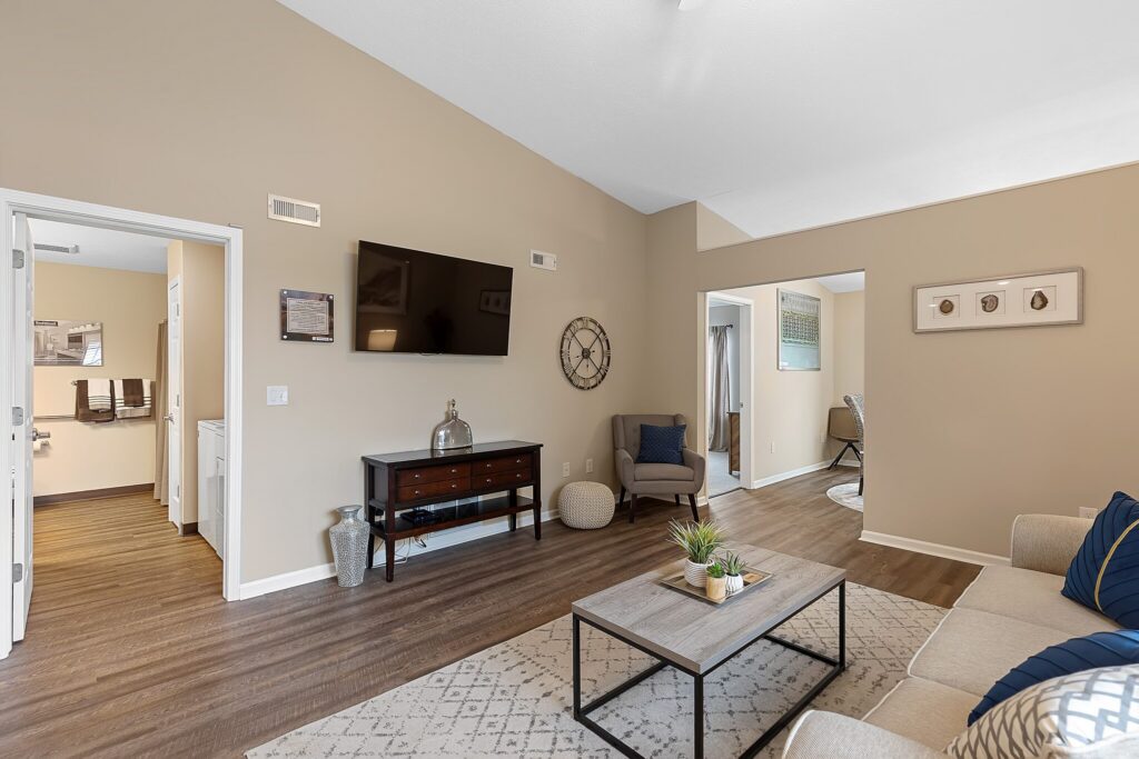 A modern living room with a wall-mounted TV, beige walls, wooden console, coffee table, sofa, and an open view into a bathroom and another room.