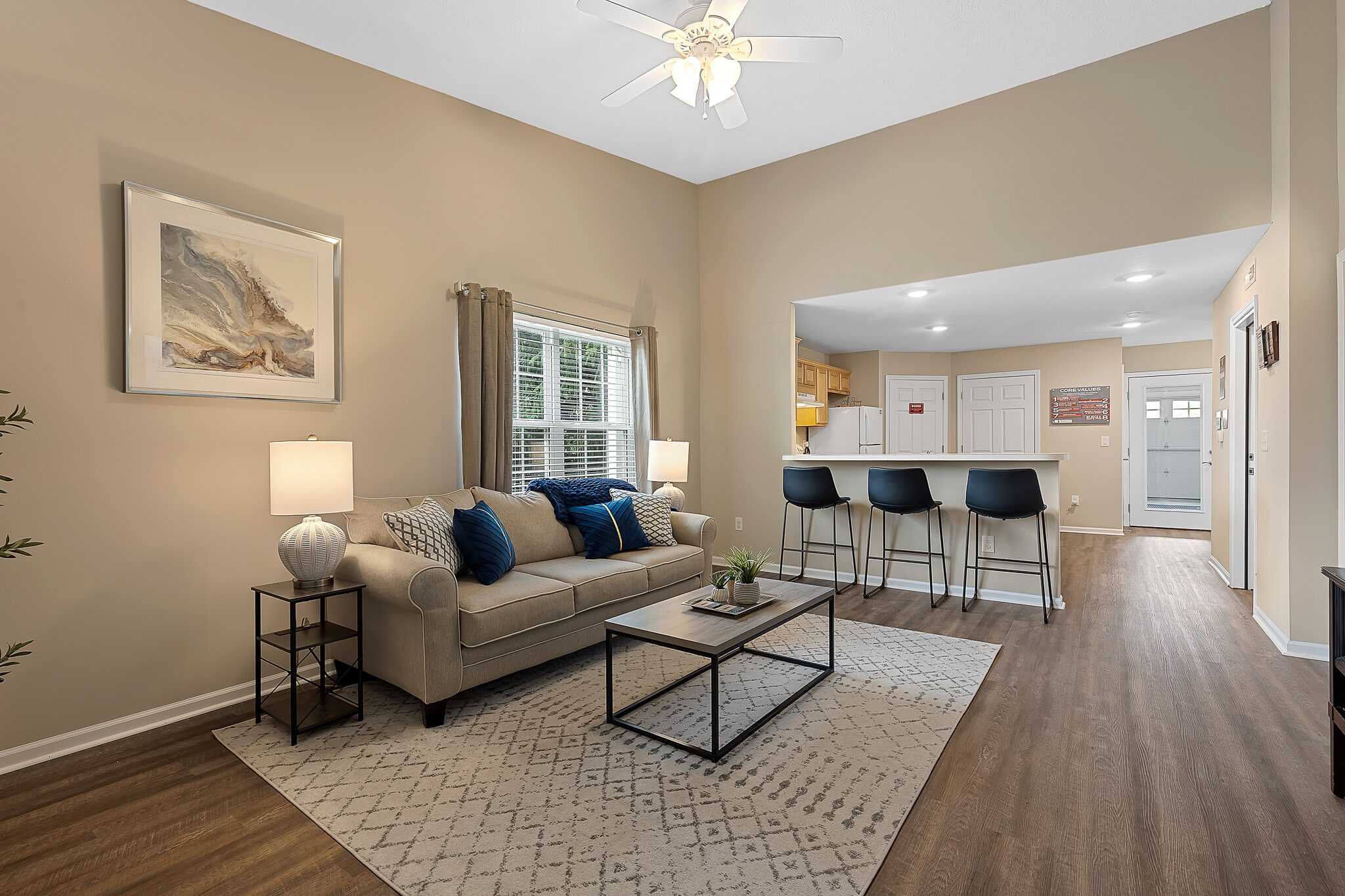 A modern living room with a beige sofa, blue accent pillows, a coffee table, and a rug, open to a kitchen with bar stools and wood flooring throughout the space.