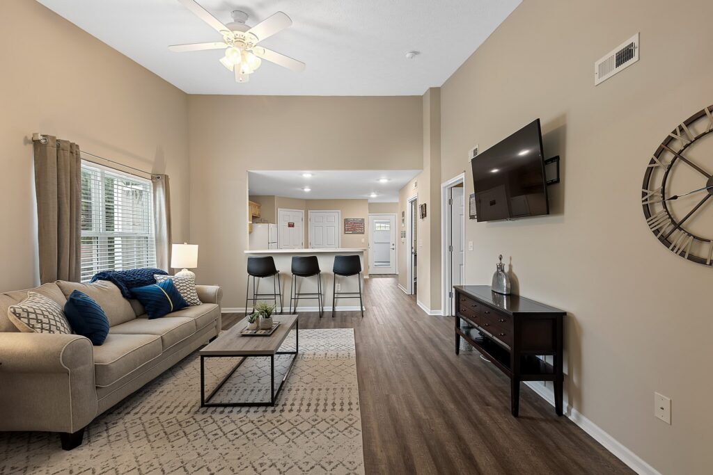 A modern living room with a beige sofa, blue pillows, coffee table, wall-mounted TV, large clock, and a breakfast bar with three black stools facing an open kitchen area.