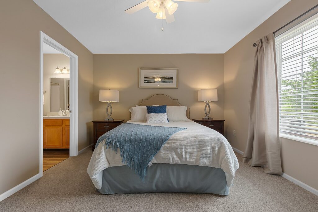 Bedroom with a neatly made bed, two bedside tables with lamps, a window with blinds and curtains, and an open door to a bathroom.
