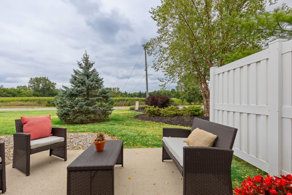 Outdoor patio with wicker furniture, beige cushions, a small table with a potted plant, and a white privacy fence, overlooking a grassy yard and trees under a cloudy sky.