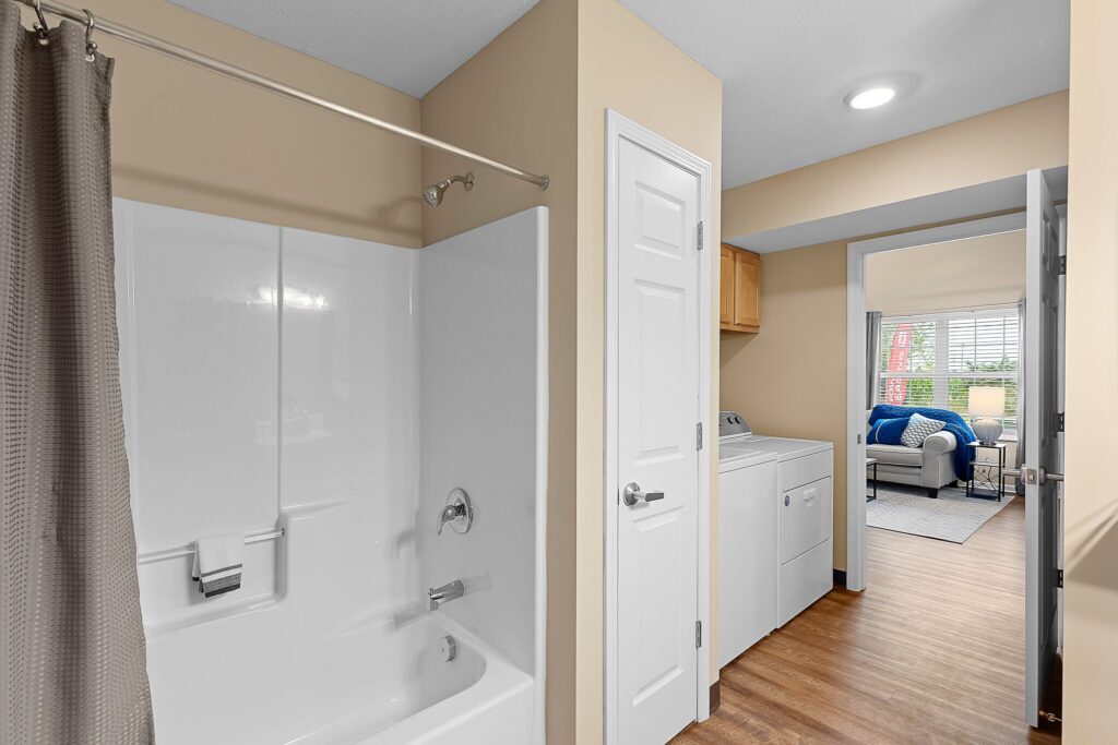 Bathroom with a white bathtub and shower combo, adjacent to a laundry area with a washer and dryer; a living room with blue accents is visible in the background.