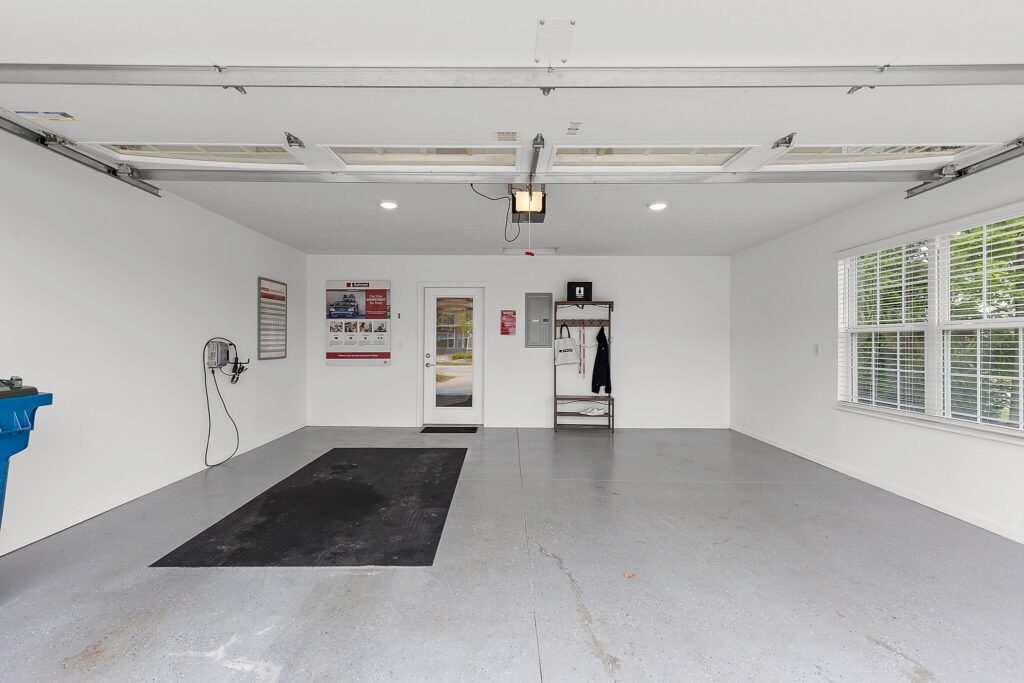 Clean, empty garage with white walls, gray floor, a large window, a door at the back, wall-mounted hose, and a small shelving unit with coats and bins.