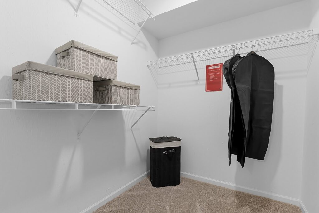 Walk-in closet with wire shelves holding three storage boxes, two garment bags hanging, a laundry hamper, and a red sign on the wall. The floor is carpeted.