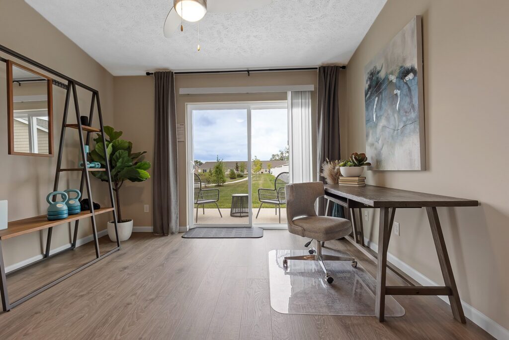 Modern home office with wood floor, desk, chair, large abstract painting, shelving unit, and potted plants. Glass doors open to a patio with outdoor chairs and green view.