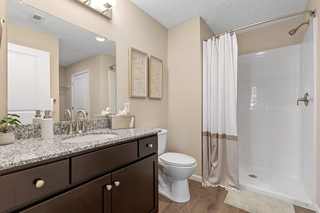 Modern bathroom with a granite countertop sink, dark wood cabinets, toilet, shower with white tiles and striped curtain, beige walls, and decorative wall art.