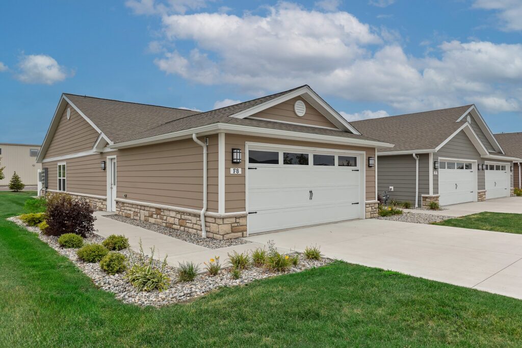Single-story suburban house with tan siding, stone accents, and a two-car garage, surrounded by a manicured lawn and landscaping under a partly cloudy sky.