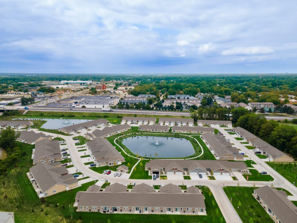 Aerial view of a suburban residential complex with a central pond and fountain, surrounded by roads, houses, and green trees under a cloudy sky.