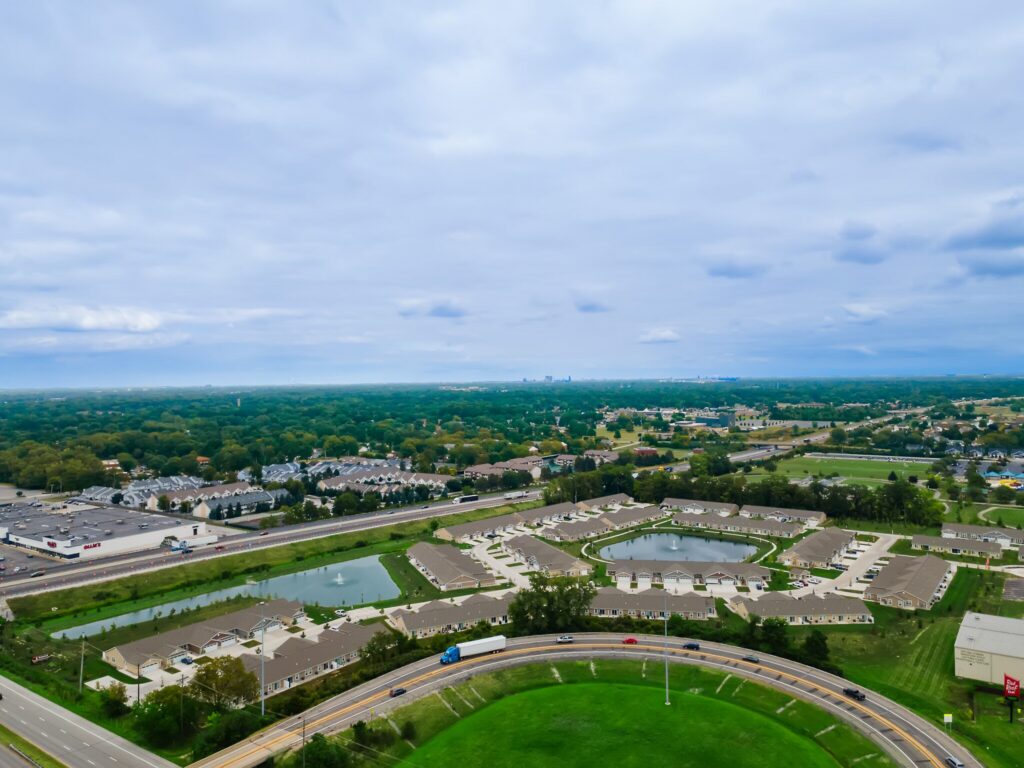 Aerial view of a suburban area with residential buildings, ponds, a curved highway, commercial properties, and a forested area in the distance under a cloudy sky.
