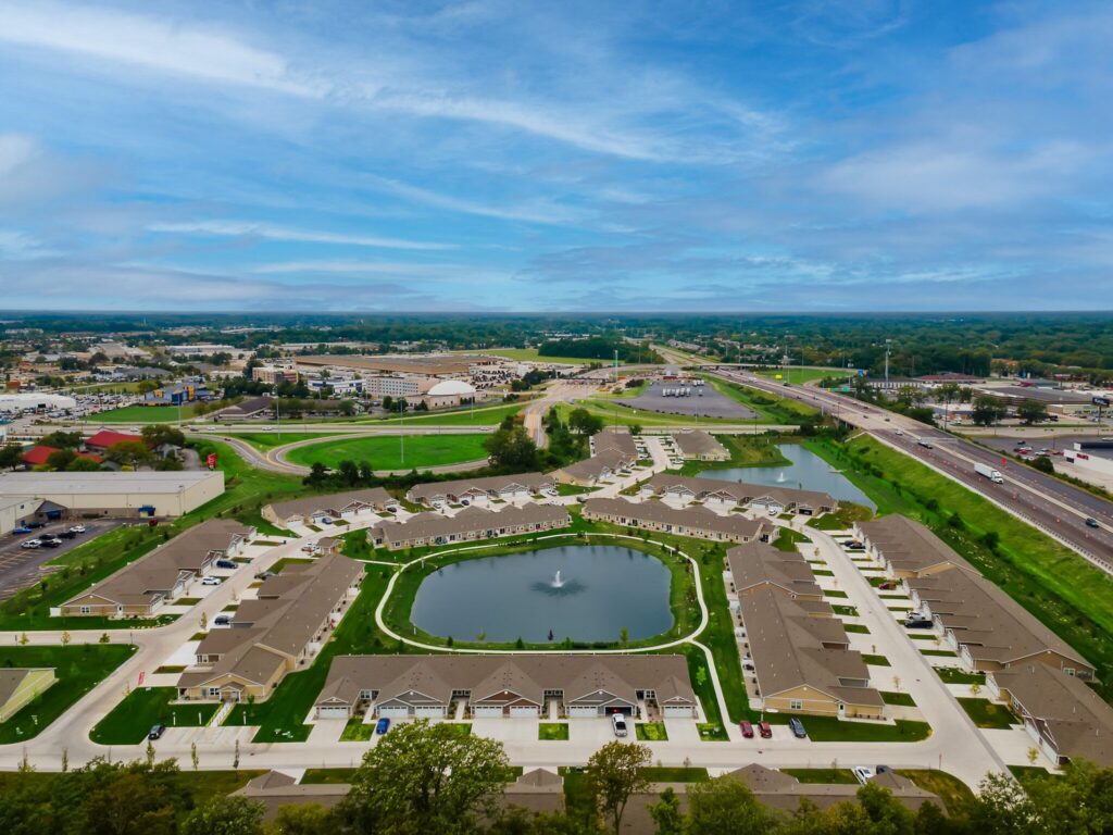 Aerial view of a residential neighborhood with houses arranged around a central pond with a fountain, surrounded by roads, green areas, and nearby commercial buildings.