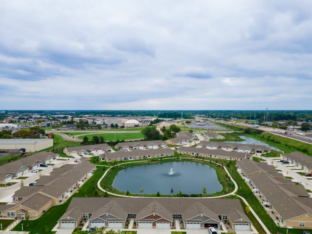 Aerial view of a suburban neighborhood with rows of houses surrounding a pond with a fountain, roads, and a commercial area in the background under a cloudy sky.