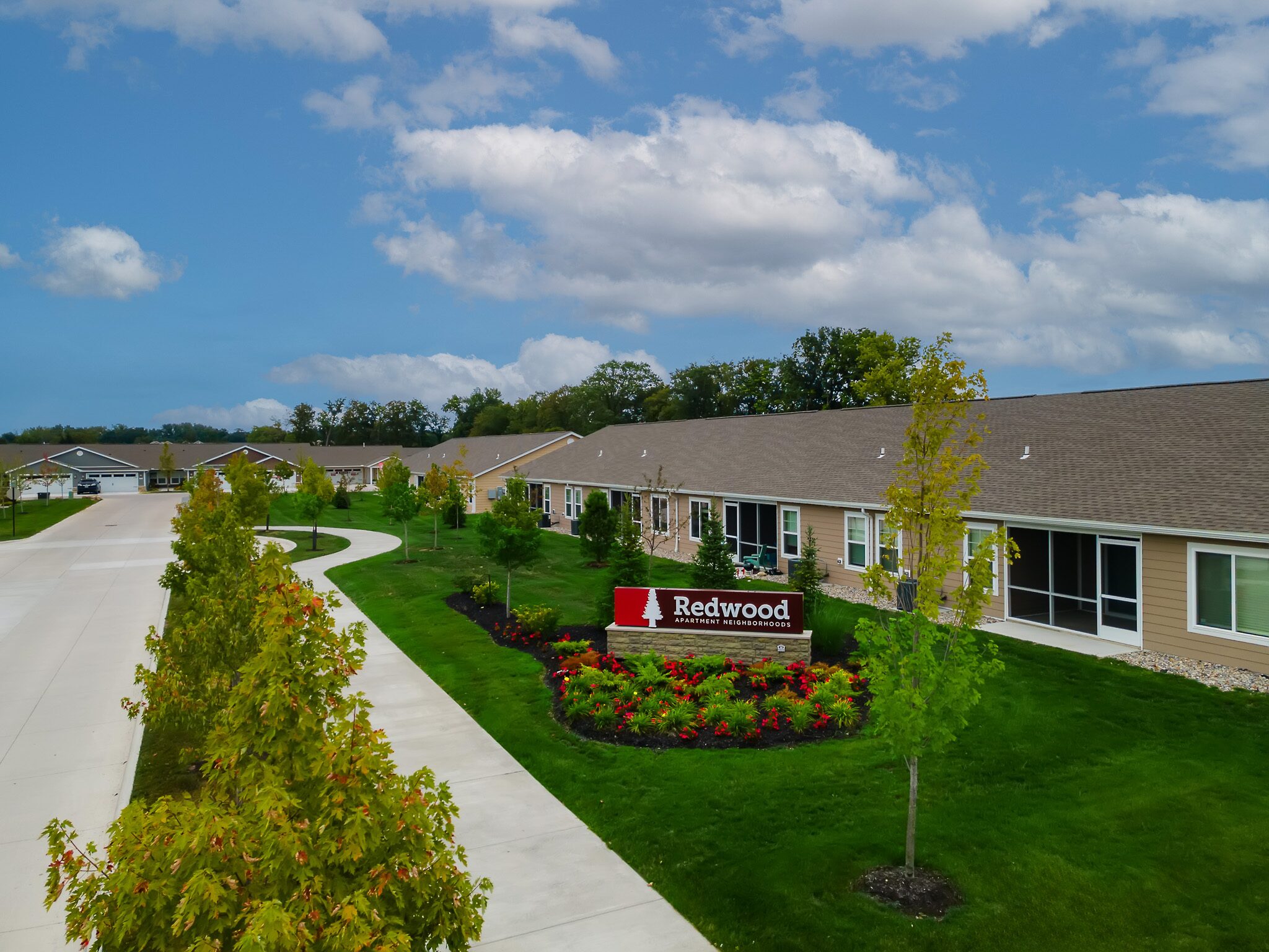 A row of single-story residential buildings with neatly landscaped lawns and a sign reading "Redwood" in front, under a partly cloudy sky.
