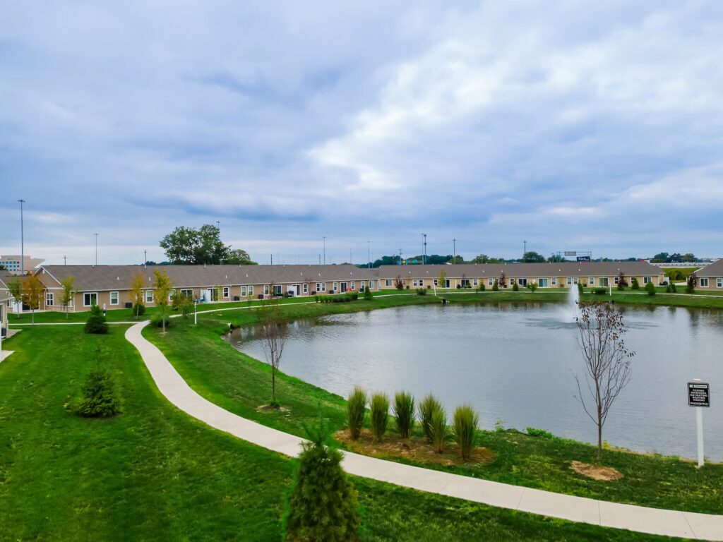 A landscaped residential area with single-story houses surrounding a pond, sidewalks, grass lawns, and a small fountain in the water under a cloudy sky.