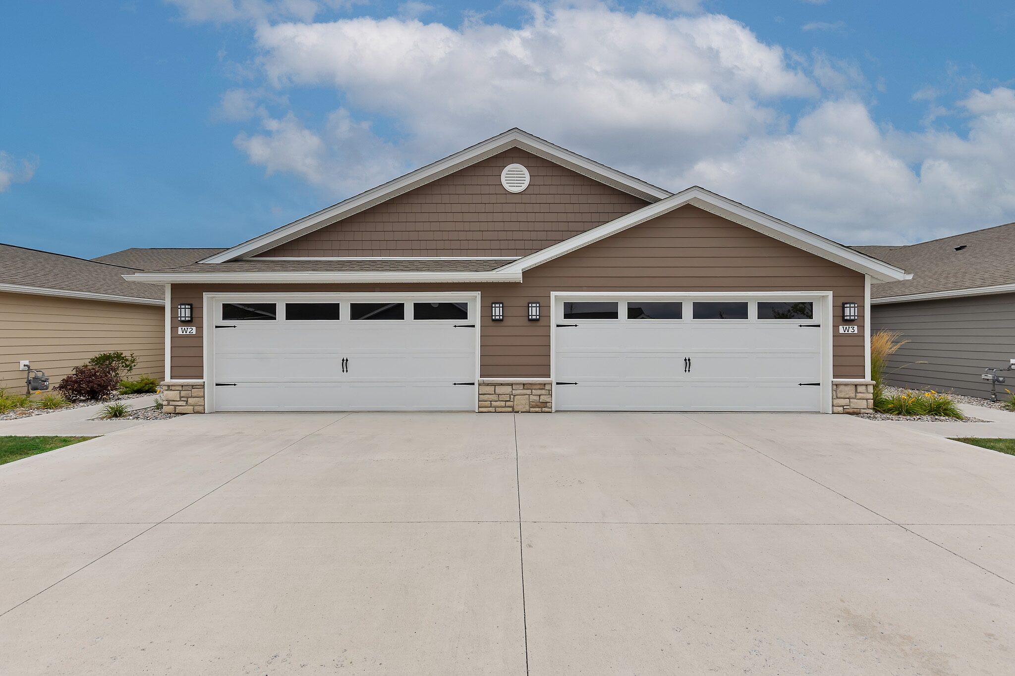 A modern duplex with two white double garage doors, brown siding, stone accents, and a wide paved driveway under a partly cloudy sky.