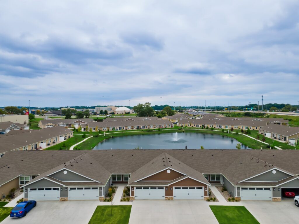 Aerial view of a suburban neighborhood with rows of attached houses facing a central pond with a small fountain, under a cloudy sky.