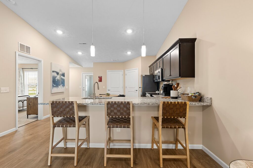 Modern kitchen with granite countertops, three woven barstools, dark cabinets, and stainless steel appliances, viewed from the dining area with a hallway and bedroom visible in the background.
