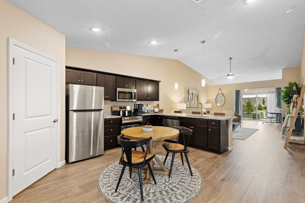 Modern kitchen with dark cabinets, stainless steel appliances, a round dining table with four chairs, and wood flooring, leading to a bright living area with large windows.