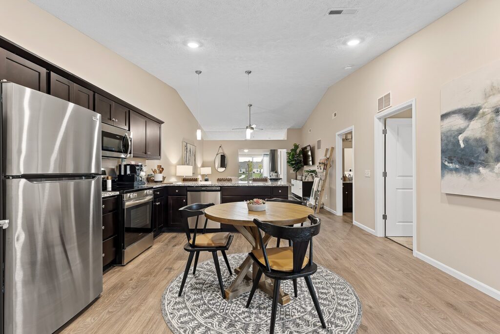 Modern kitchen and dining area with stainless steel appliances, dark cabinets, round wooden table with four chairs, and neutral decor on wood flooring.