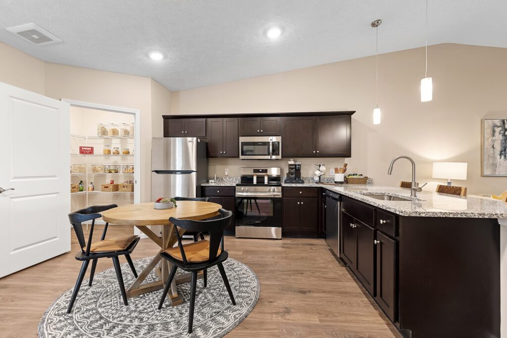 Modern kitchen with dark cabinets, stainless steel appliances, a round dining table with four chairs, and an open pantry stocked with jars and containers.
