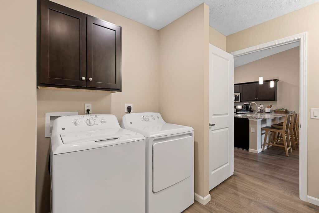A laundry room with a white washing machine and dryer beneath dark cabinets, with a view into a kitchen and dining area through an open doorway.