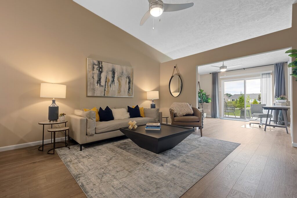 Modern living room with beige walls, neutral furniture, abstract art, and a large window showing a balcony with greenery outside. Ceiling fan and lamps provide lighting.