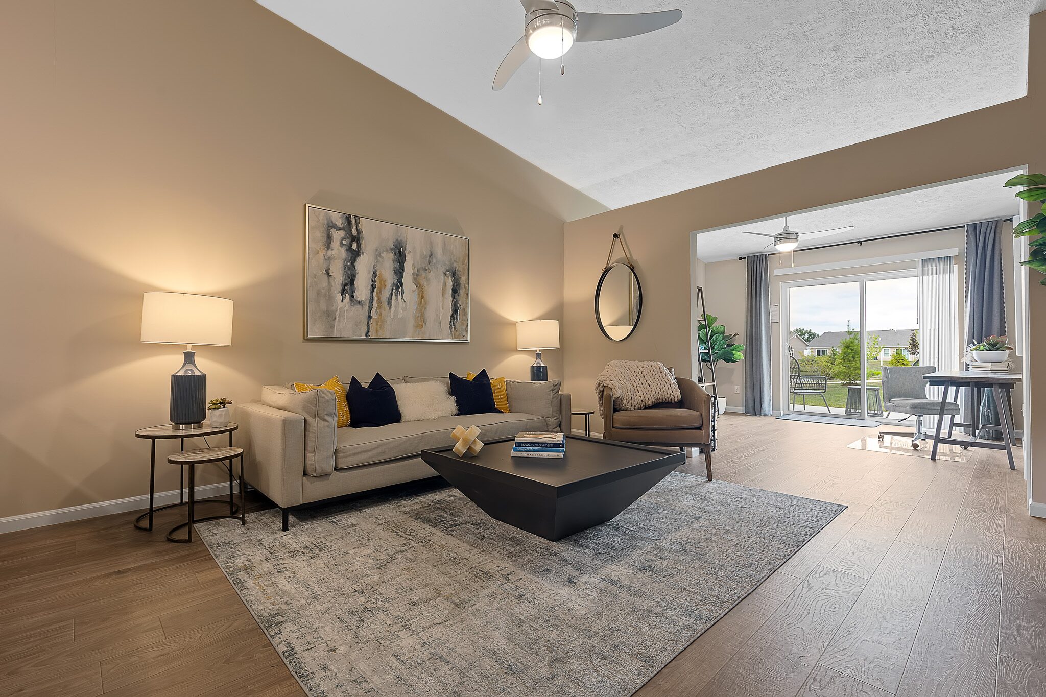 Modern living room with beige walls, neutral furniture, abstract art, and a large window showing a balcony with greenery outside. Ceiling fan and lamps provide lighting.