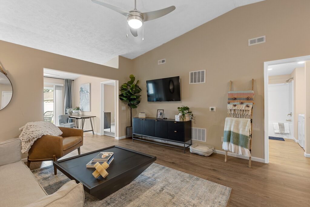 Modern living room with neutral tones, a ceiling fan, wall-mounted TV, contemporary furniture, a large plant, and an adjoining view into a bathroom and another room.