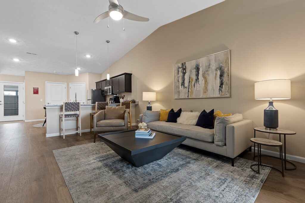 Modern living room with a beige sofa, abstract wall art, and a large coffee table. Kitchen with barstools and pendant lights visible in the background. Neutral color palette throughout.
