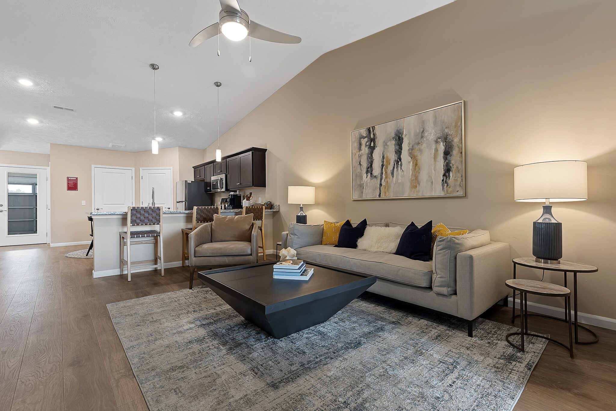 Modern living room with a beige sofa, abstract wall art, and a large coffee table. Kitchen with barstools and pendant lights visible in the background. Neutral color palette throughout.