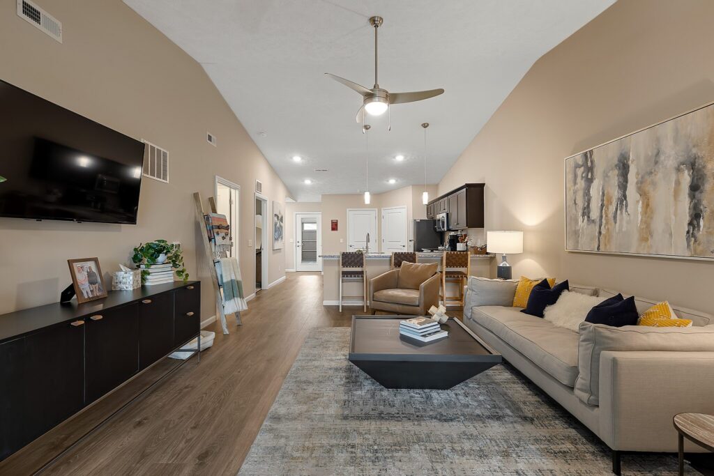 Modern living room with beige walls, a mounted TV, a gray sofa, abstract art, and an open layout leading to the kitchen and dining area.