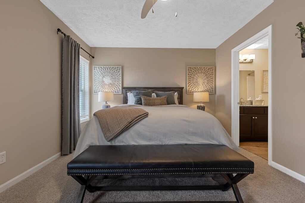 A neatly made bed with gray bedding, two bedside tables with lamps, a black bench at the foot, and an adjoining bathroom visible through an open door.